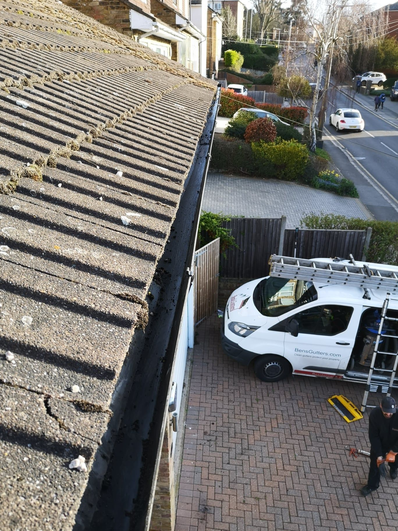 White van in a driveway below a rooftop view, with a person standing beside it and a suburban street beyond.
