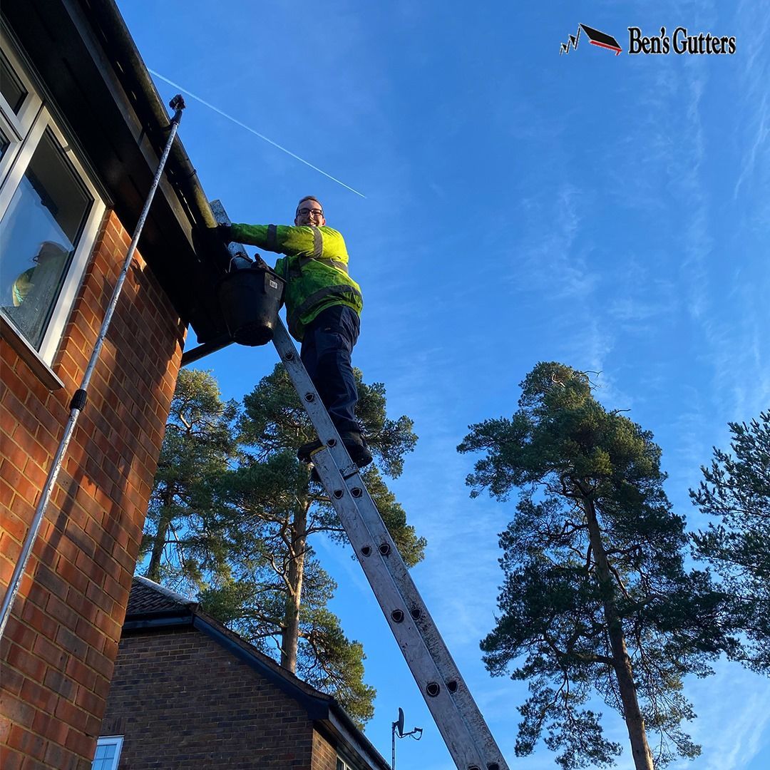 Worker on a ladder cleaning a brick house roofline against a blue sky