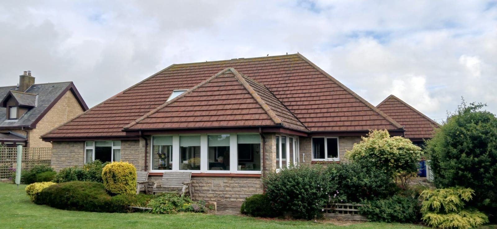 Single-story house with red-tiled roof, bay windows, and a green garden under a cloudy sky