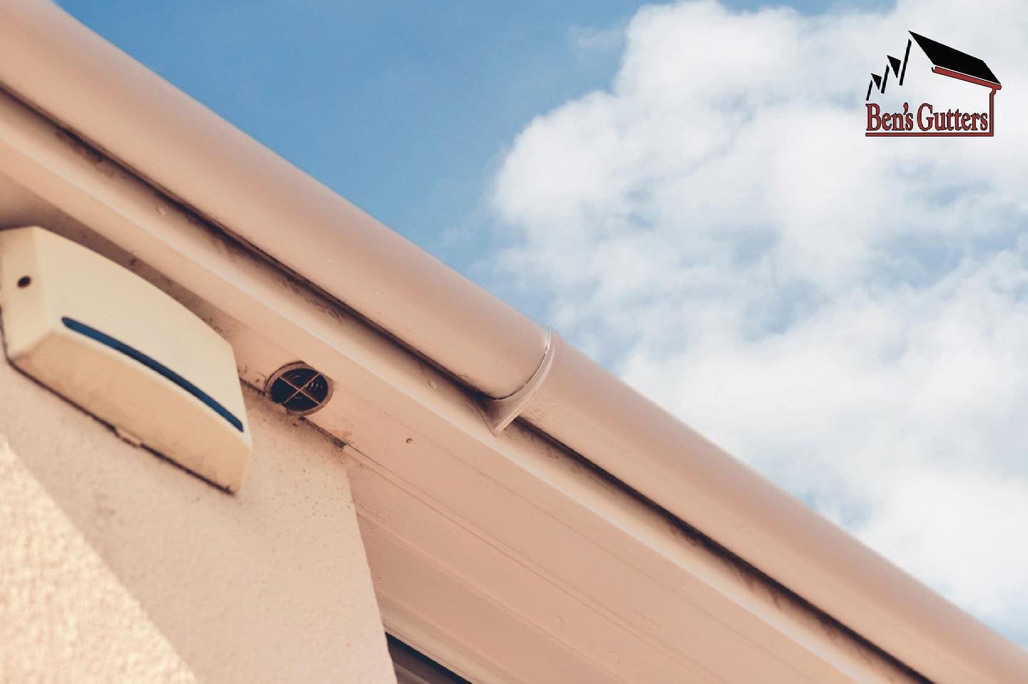 Beige rain gutter against a blue sky with clouds; a rectangular white box attached to the wall.