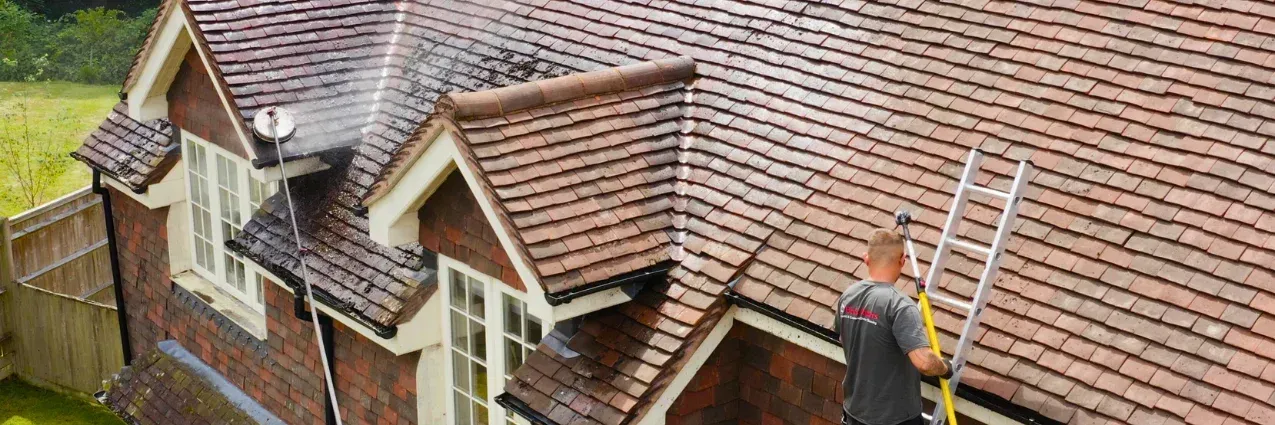 Worker on a roof with a ladder beside a brick house and tiled roof