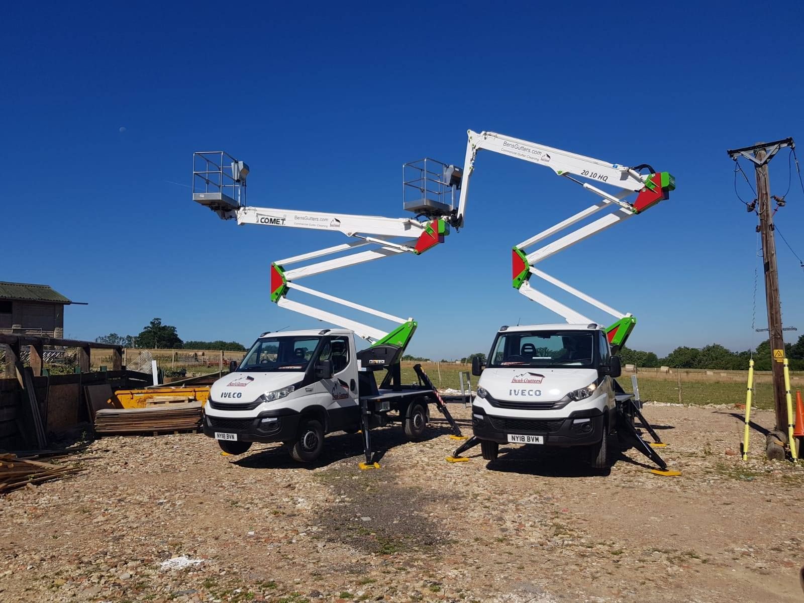 Two white utility trucks with raised cherry picker booms in a gravel lot under a clear blue sky