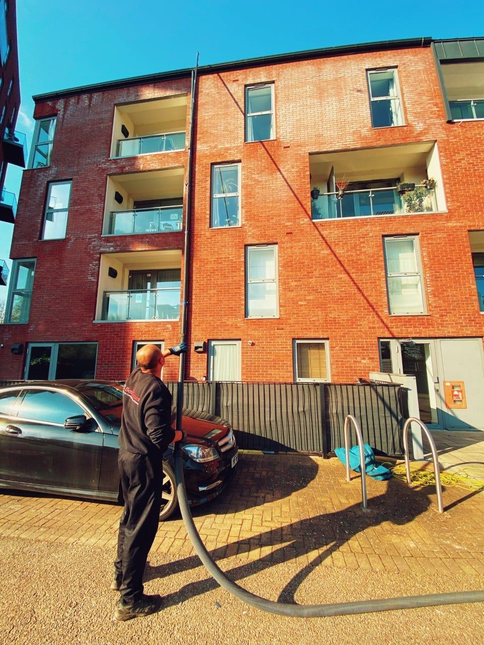 Person stands outside a red brick apartment building near a parked car and a bicycle.