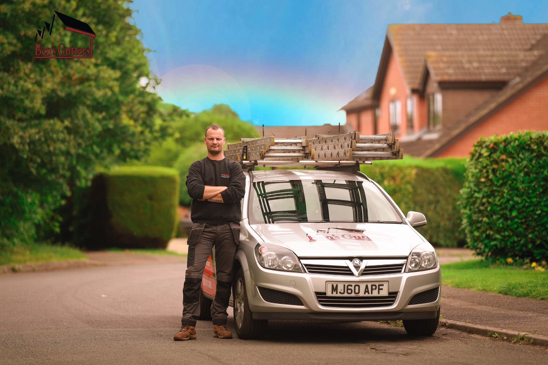 Man standing beside a silver car with ladders on the roof, parked on a suburban street under a rainbow.