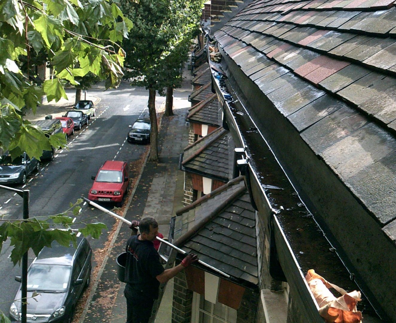 Person on a rooftop walkway beside parked cars and trees along a street below.