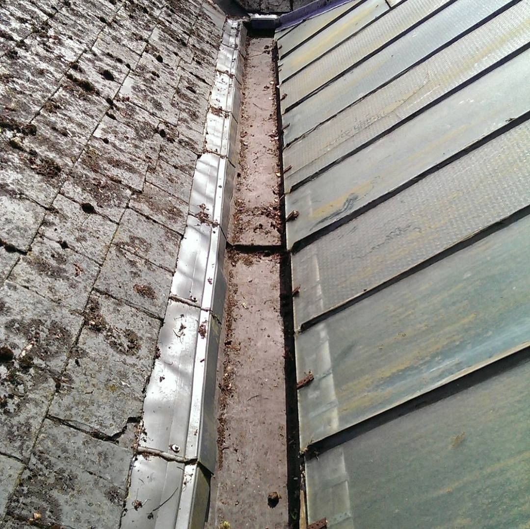 Roof gutter between a brick wall and metal roof panels, showing dirt and debris buildup.
