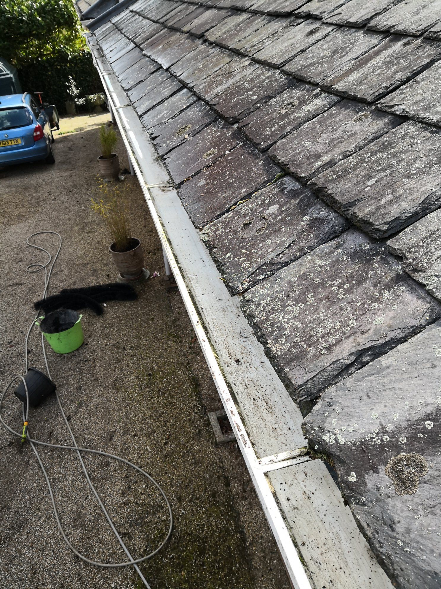 Rain gutter along a shingled roof over a gravel driveway with a blue car and scattered tools below