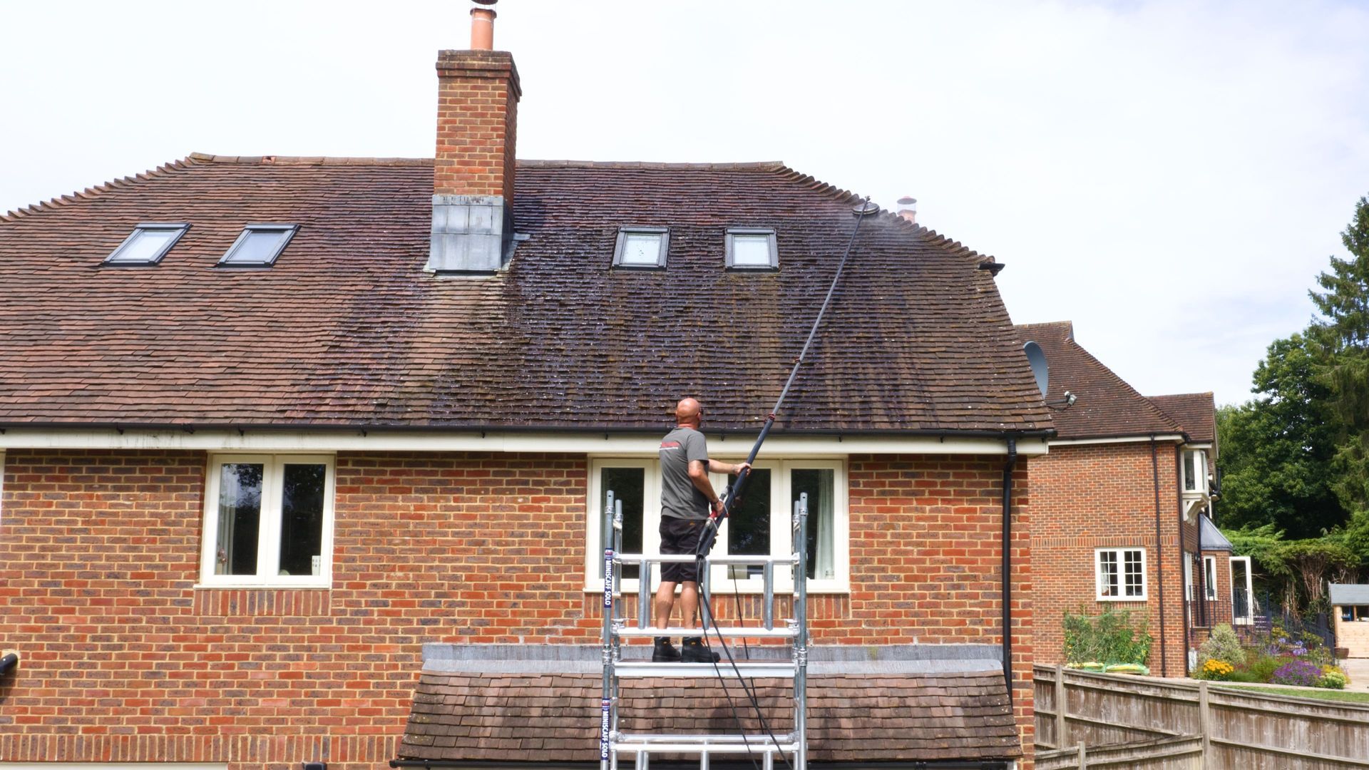 Worker on scaffold painting a brick house exterior beneath skylights and a chimney.
