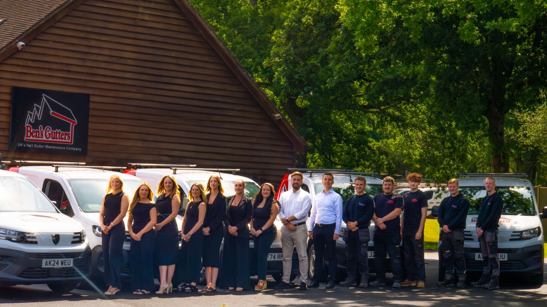 Group of people standing beside white vans and a black car in front of a wooden building outdoors
