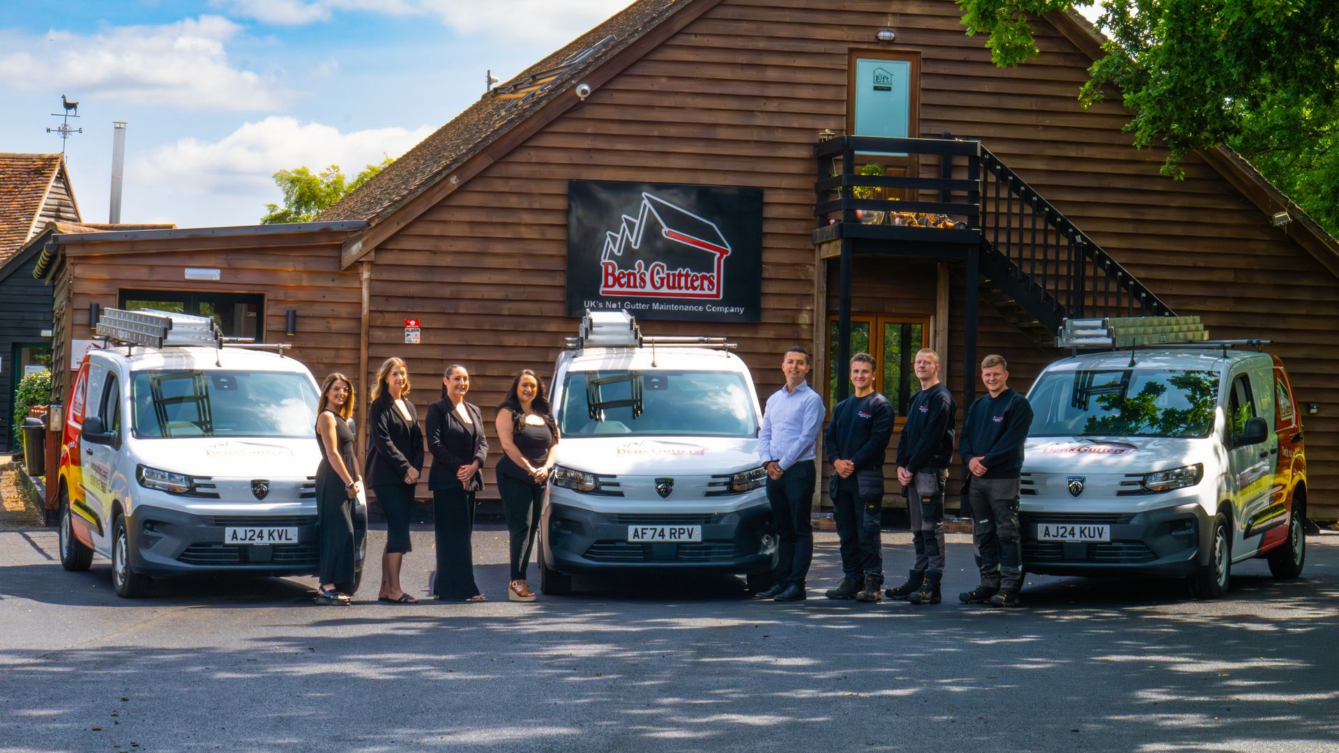 Group posed in front of three white vans outside a wooden building with a sign and flag.