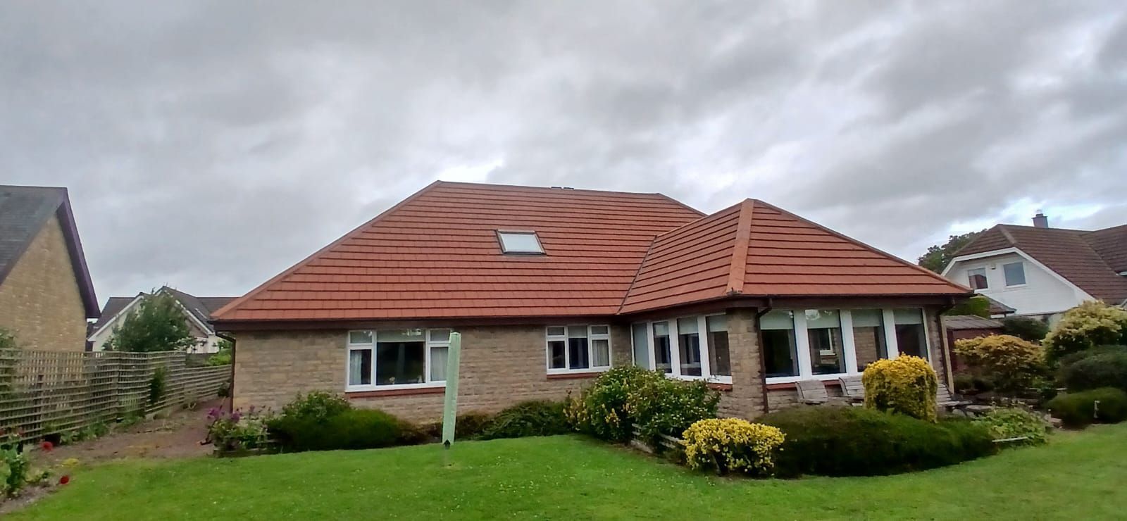 Single-story brick house with red tiled roof, green lawn, and shrubs under a cloudy sky