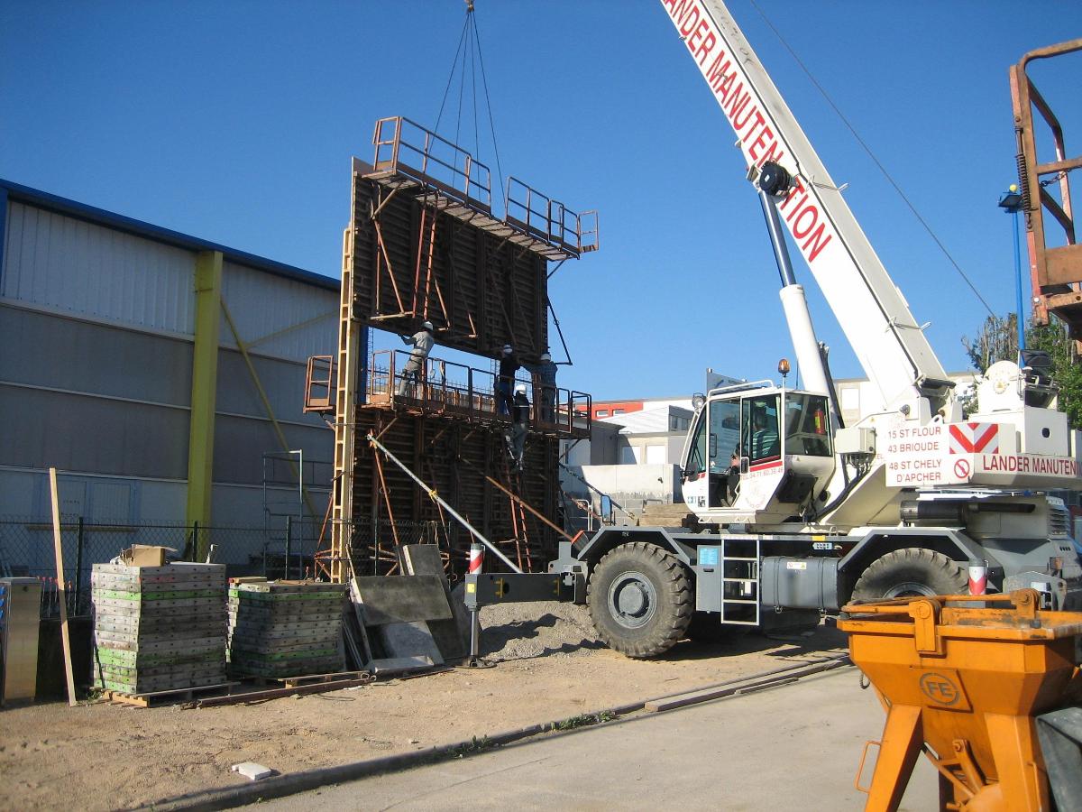 Une grue soulève un grand panneau de coffrage brun sur un chantier de construction par une journée ensoleillée.
