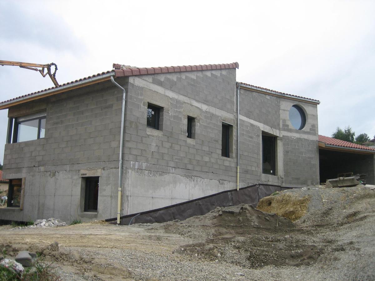 Construction d'une maison à deux étages en parpaings de béton gris avec un toit de tuiles rouges, entourée de terre et de gravier.