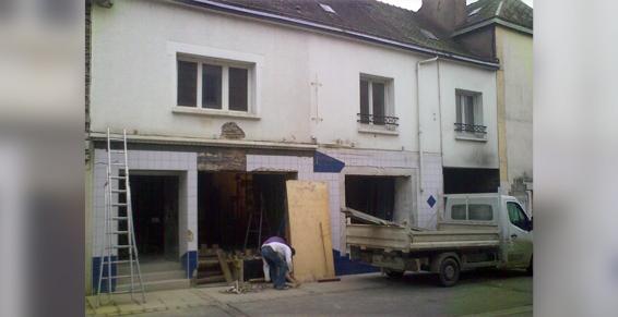 Extérieur du bâtiment en rénovation; façade blanche, ouvriers, camion, bois et échafaudages présents.