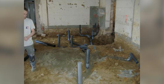 Un homme en bottes se tient près de tuyaux de plomberie exposés sur un chantier de construction ; sol en terre battue.