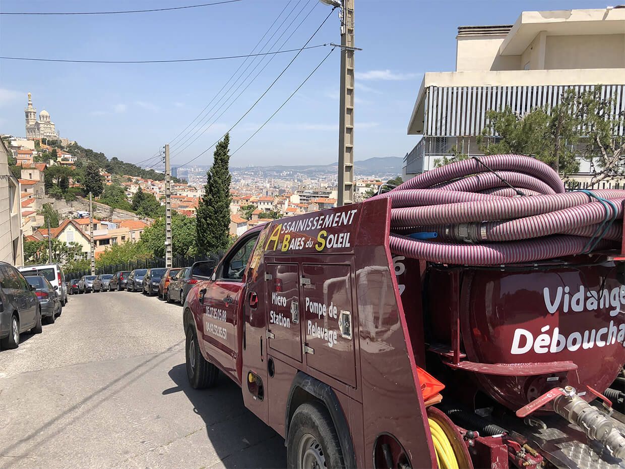 Camionnette bordeaux d'assainissement transportant des tuyaux enroulés sur une rue escarpée surplombant une ville
