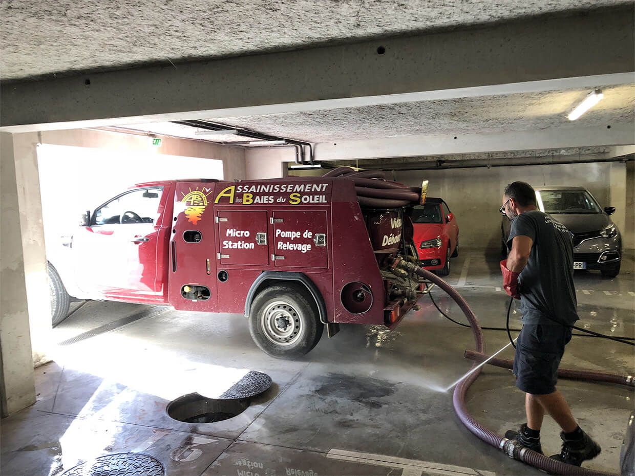 Camion d'assainissement rouge et ouvrier dans un parking souterrain en béton, avec un tuyau d'arrosage au sol.