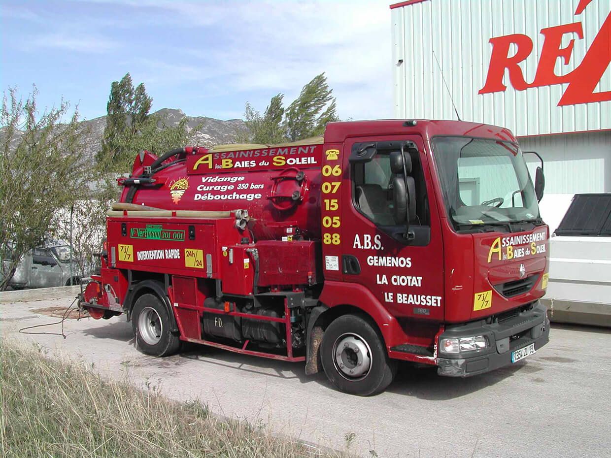 Camion d'assainissement rouge à bras hydraulique stationné en plein jour au bord d'une route et d'un bâtiment