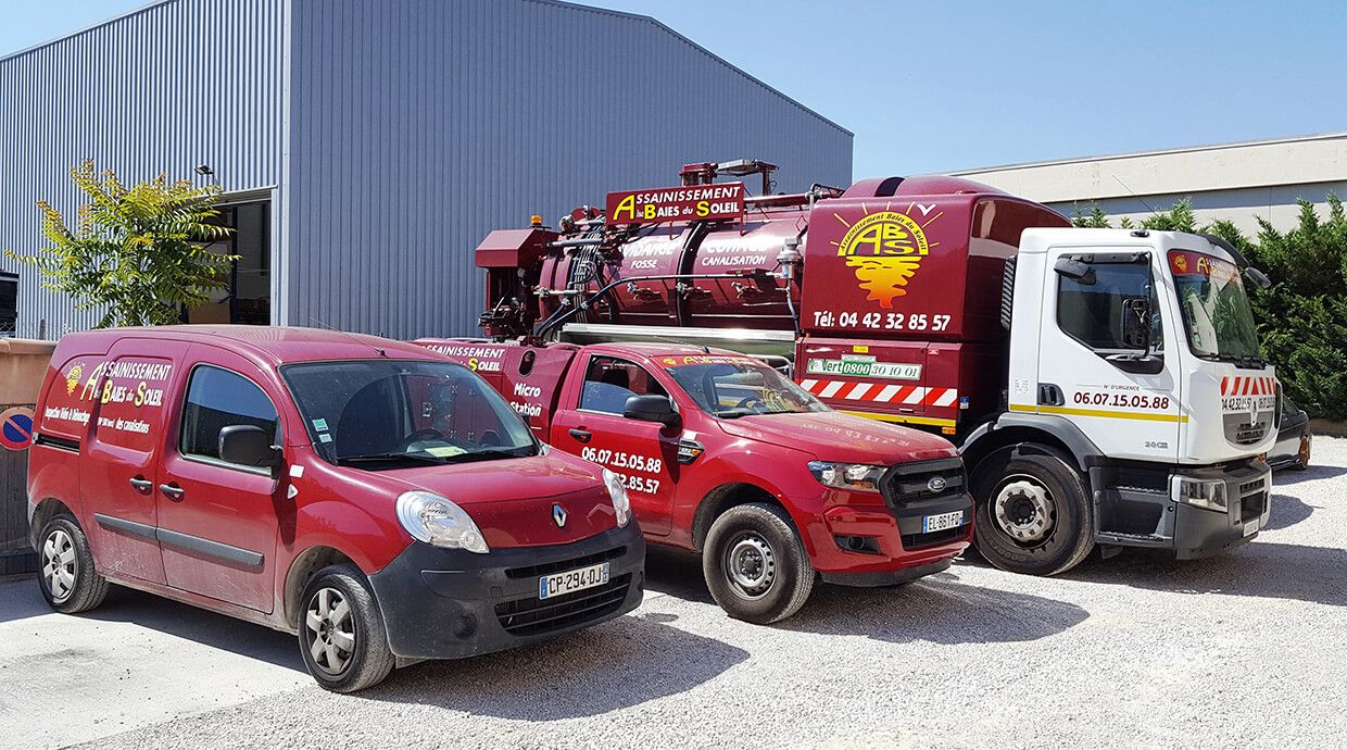 Des fourgonnettes de service rouges et un camion d'assainissement blanc sont stationnés devant un bâtiment métallique.