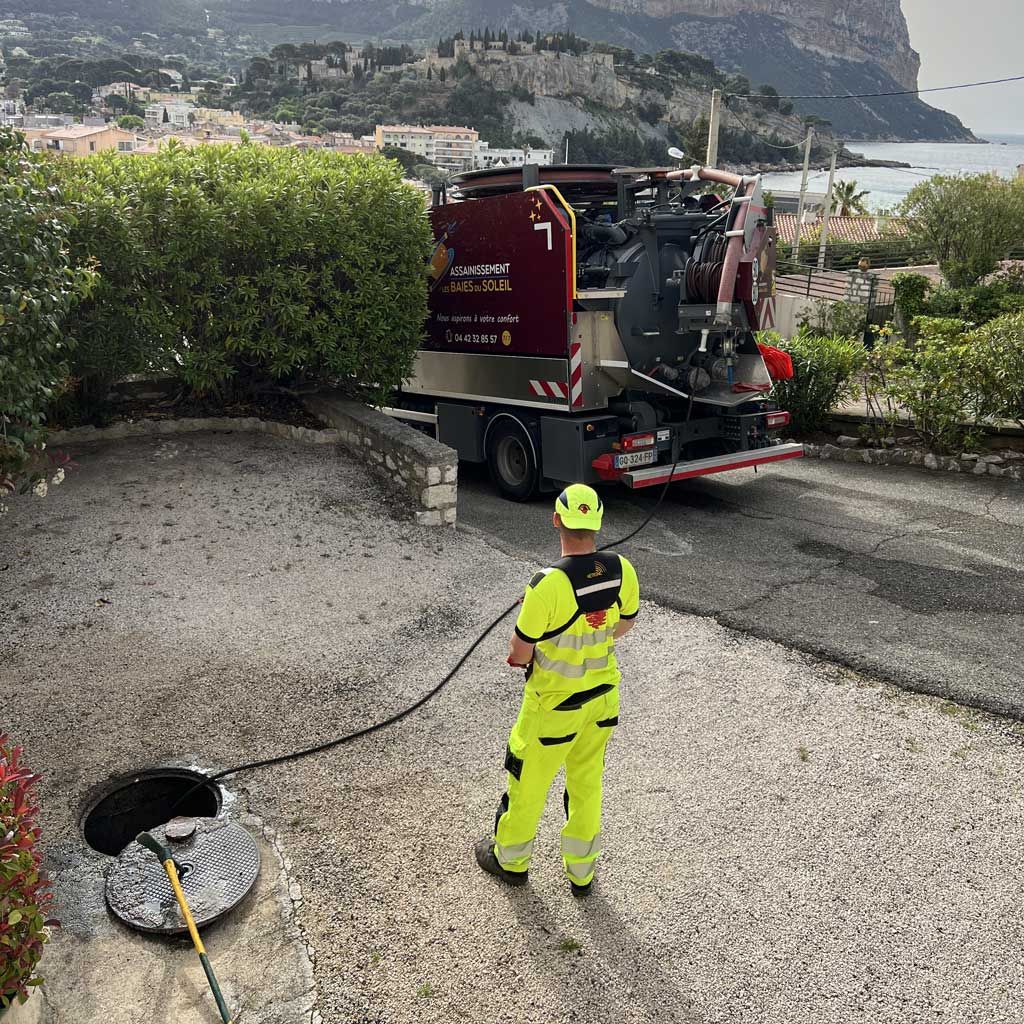 Un ouvrier vêtu d'une combinaison de sécurité fluo se tient près d'un tuyau d'évacuation des eaux pluviales, à proximité d'un camion utilitaire rouge, sur une route côtière.