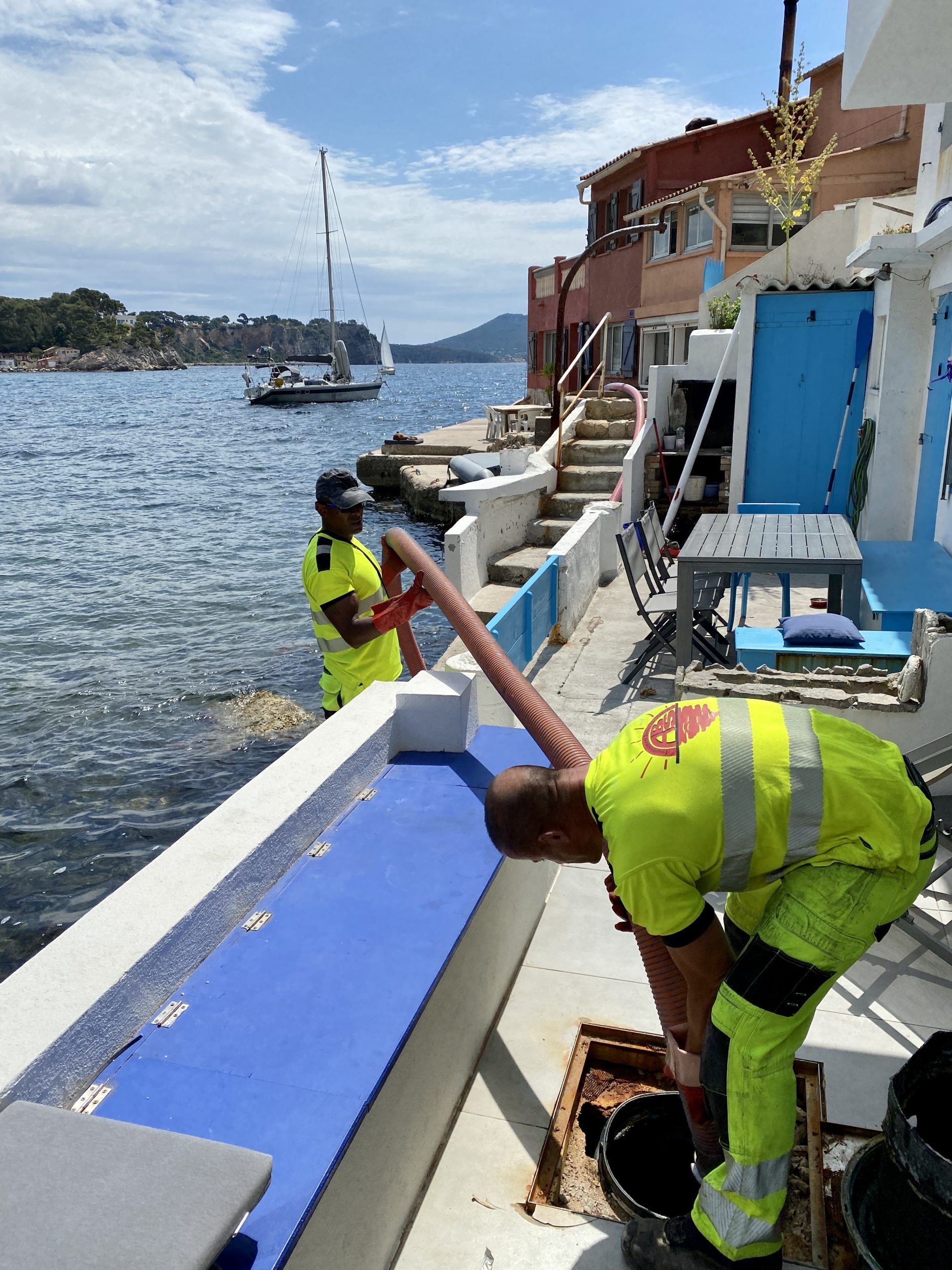 Des ouvriers vêtus d'équipements de sécurité jaunes réparent un quai au bord d'une eau bleue où sont amarrés des bateaux.