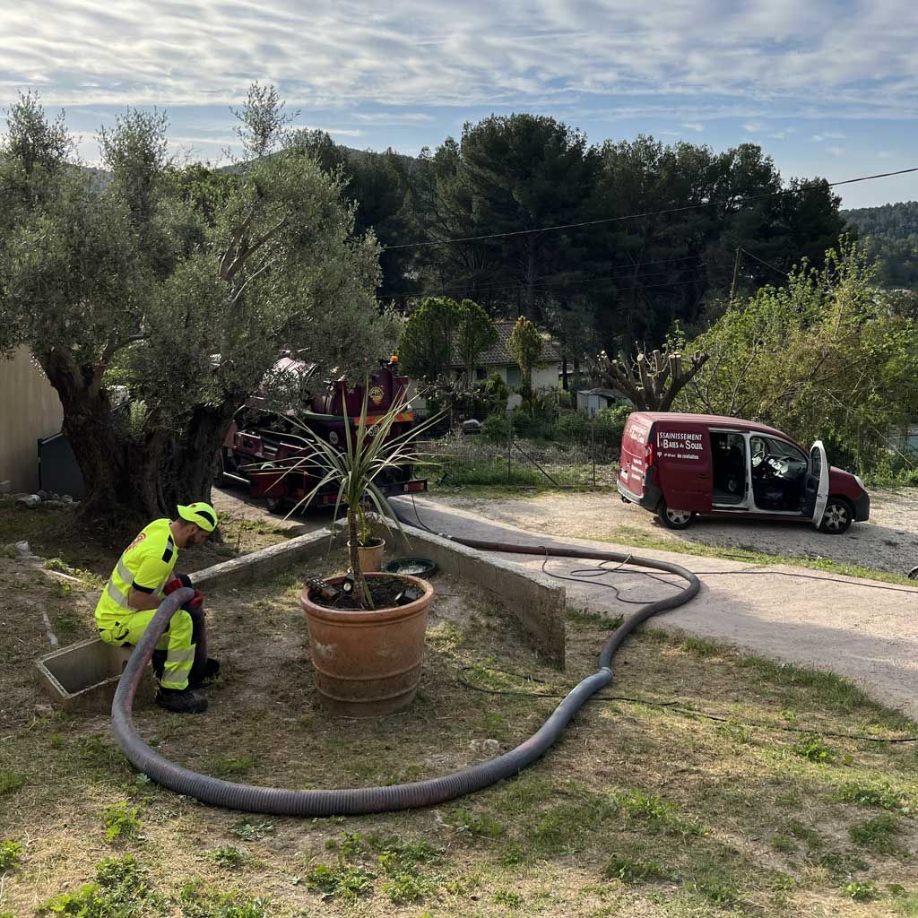 Un ouvrier avec un tuyau d'arrosage près d'une plante en pot ; une camionnette rouge garée sur une allée en pente dans une cour.