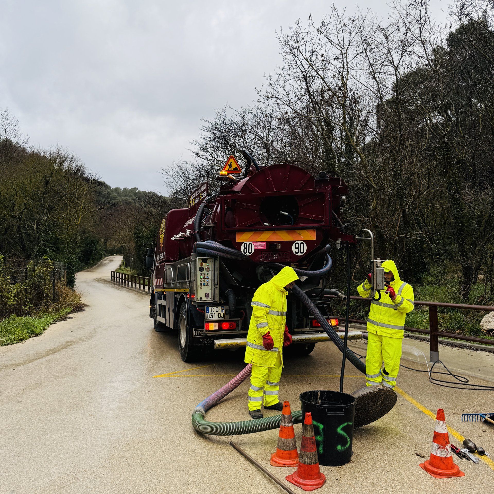 Deux ouvriers en combinaisons jaunes interviennent près d'un camion d'assainissement rouge sur le bord d'une route mouillée, avec des cônes et un tuyau d'arrosage à proximité.
