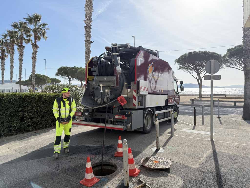 Camion d'assainissement à côté d'une bouche d'égout avec deux employés en combinaisons de sécurité jaunes sur une route côtière