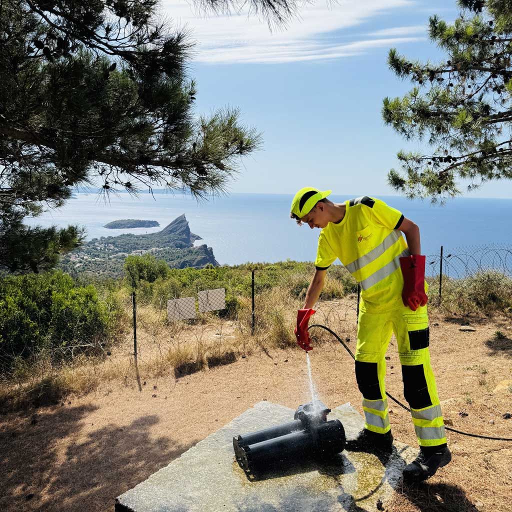 Un ouvrier en tenue de sécurité fluo utilise un tuyau d'arrosage au bord d'une route, sous un arbre, avec une vue côtière en arrière-plan.