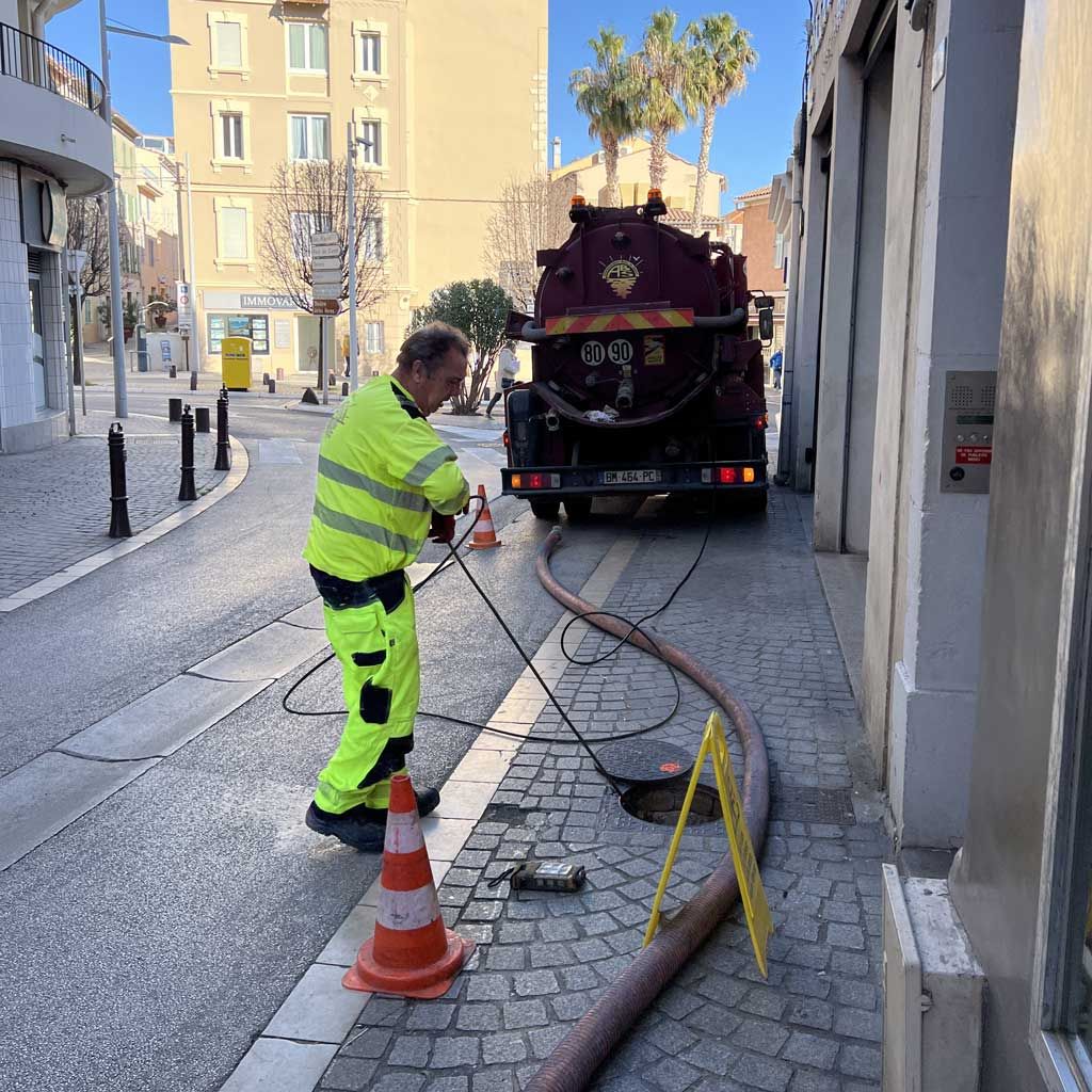 Un ouvrier en combinaison de sécurité fluorescent utilise un tuyau d'arrosage raccordé à un camion d'assainissement dans une rue étroite.