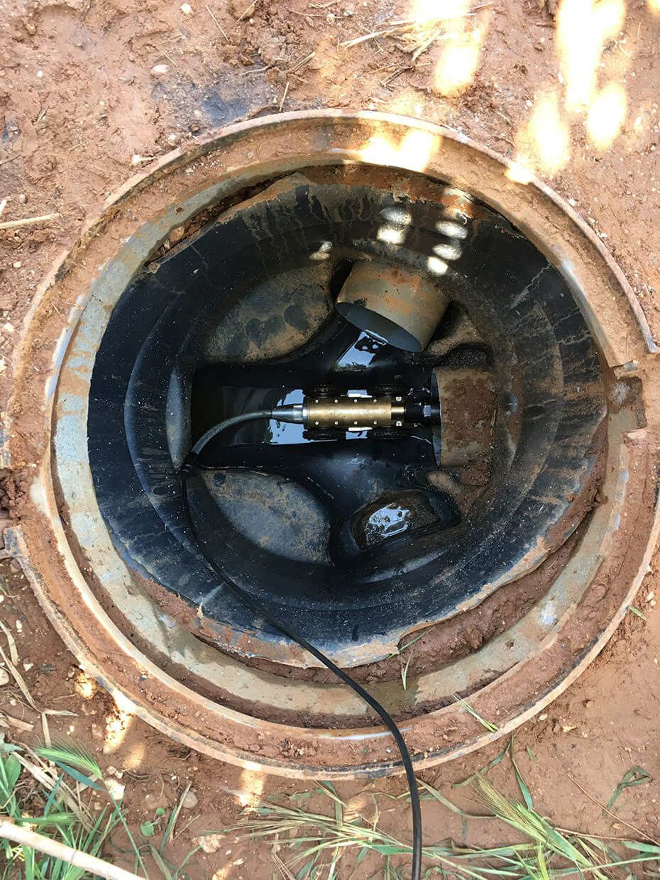 Trou d'égout ouvert avec une pompe et un tuyau noirs à l'intérieur, entouré de terre.
