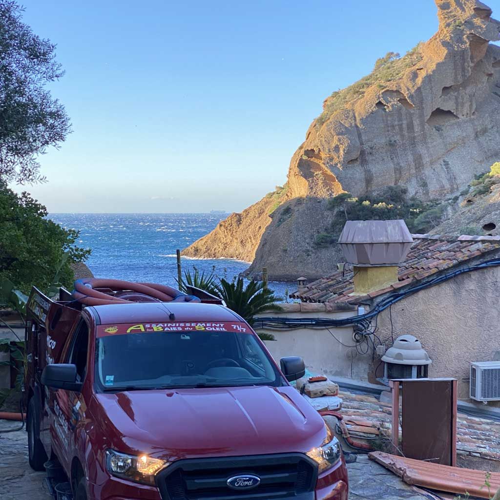 Une camionnette rouge sur une route côtière étroite, au bord des falaises, avec la mer et le ciel bleu en arrière-plan.