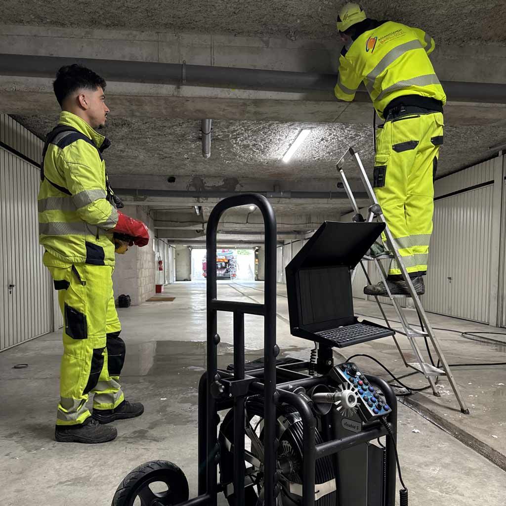 Des ouvriers en tenue de sécurité fluo inspectent le plafond en béton d'un tunnel à proximité d'un chariot et d'une échelle.
