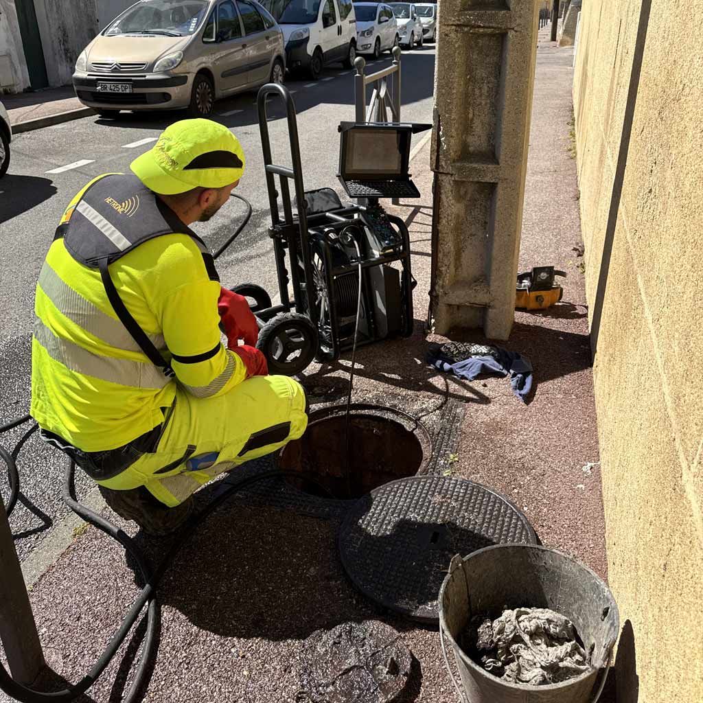 Un ouvrier en tenue de sécurité jaune inspecte une bouche d'égout ouverte en bordure de rue, du matériel se trouvant à proximité.