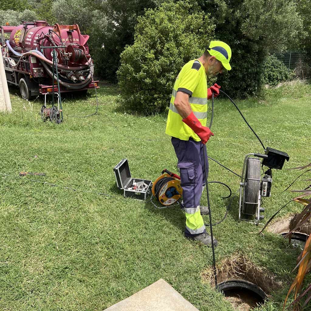 Un ouvrier portant une tenue de sécurité fluo inspecte un regard d'égout à l'aide d'équipements dans une zone herbeuse extérieure.