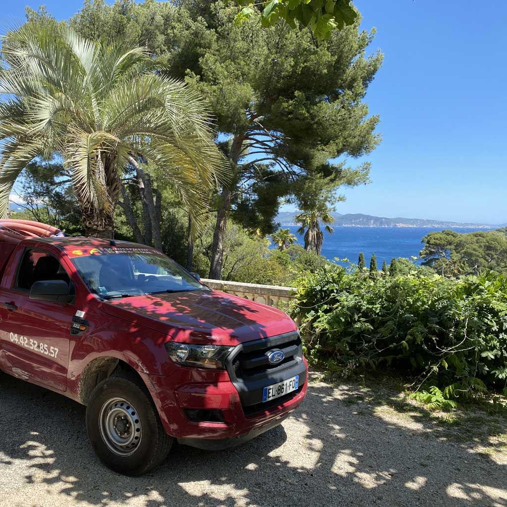 Un pick-up rouge sur une route tropicale ensoleillée, bordée de palmiers et offrant une vue sur l'océan au loin.