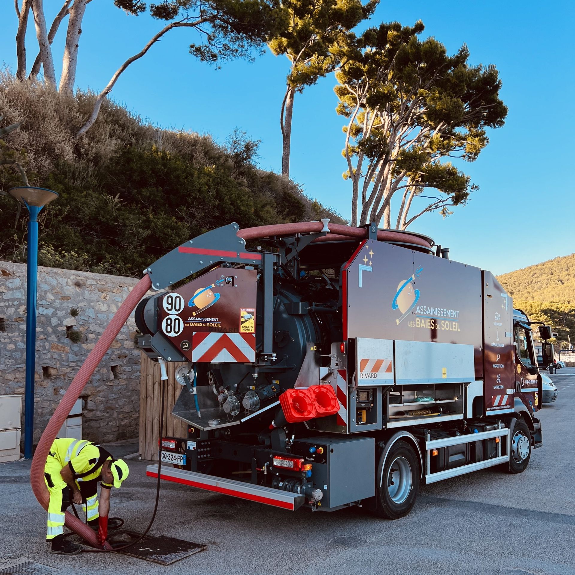 Camion d'assainissement avec un ouvrier nettoyant la chaussée à côté d'un mur de pierre et d'arbres.