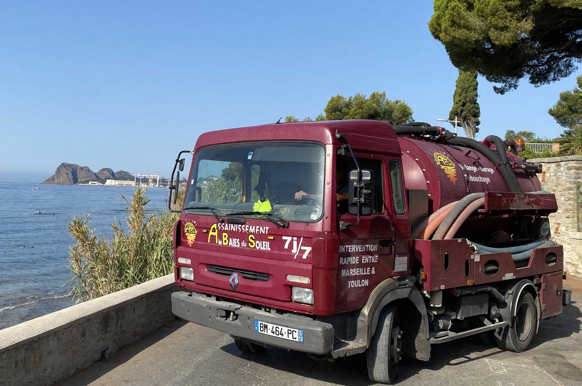 Camion balayeuse bordeaux stationné au bord de la route côtière, avec l'eau bleue et des îlots rocheux en arrière-plan.
