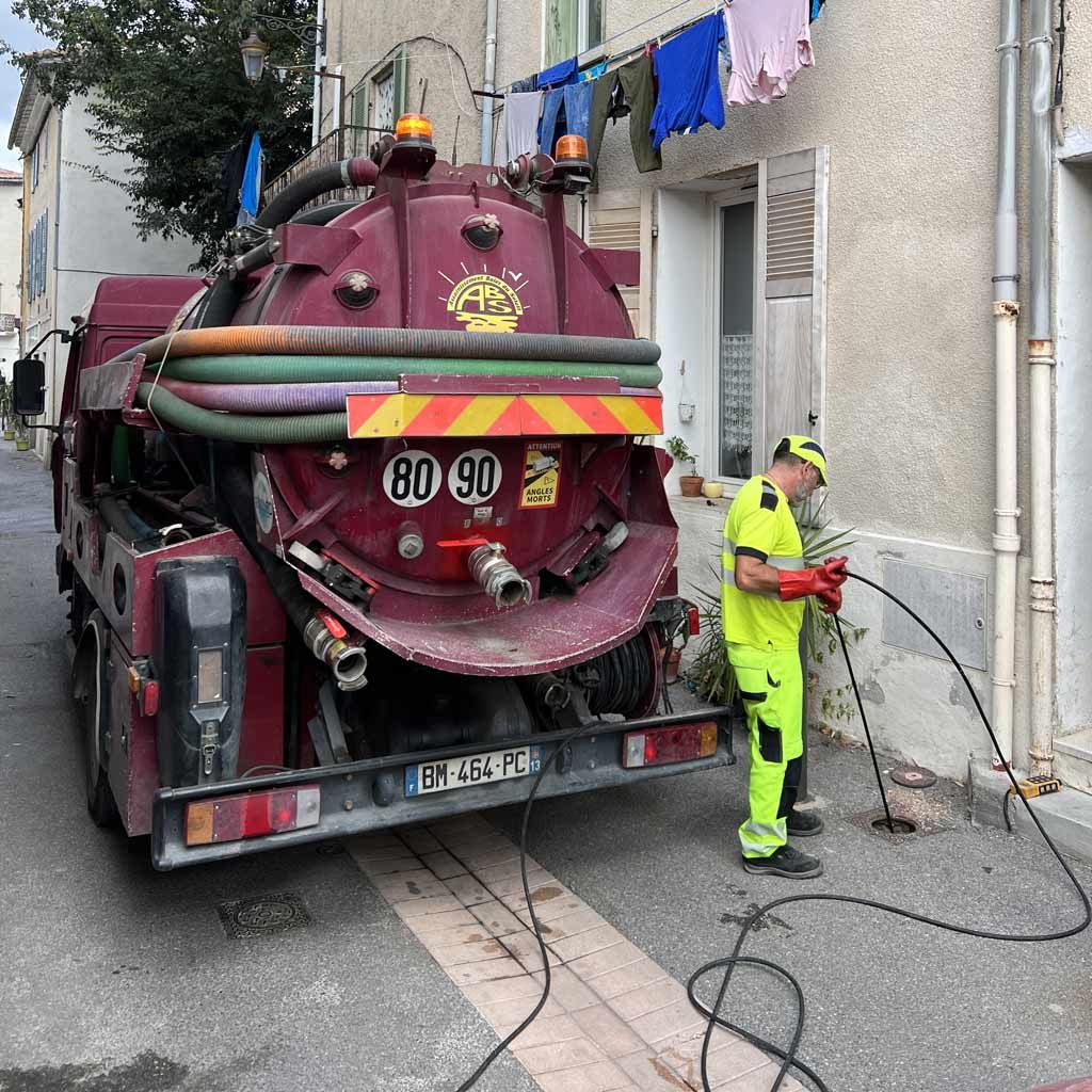 Un ouvrier vêtu de jaune fluo nettoie un camion aspirateur rouge dans une ruelle étroite, les tuyaux étant posés au sol.