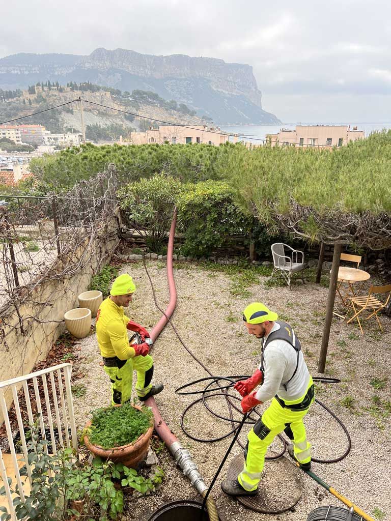 Deux ouvriers vêtus de tenues de sécurité fluo taillent des vignes dans un jardin avec des montagnes en arrière-plan.