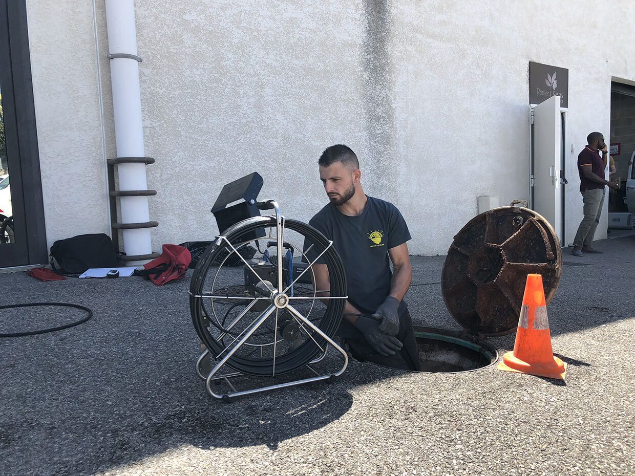 Un homme est agenouillé près d'une grande roue posée sur du gravier, à proximité d'un bâtiment ; un cône orange et des pièces de vélo se trouvent à proximité.