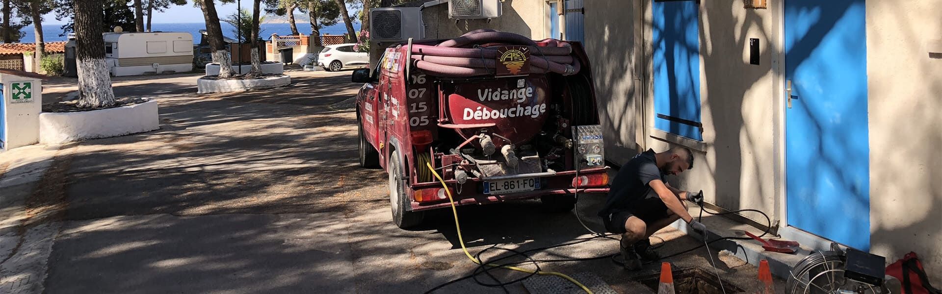 Une personne est accroupie près d'un camion d'assainissement bordeaux dans une rue ensoleillée aux murs bleus.