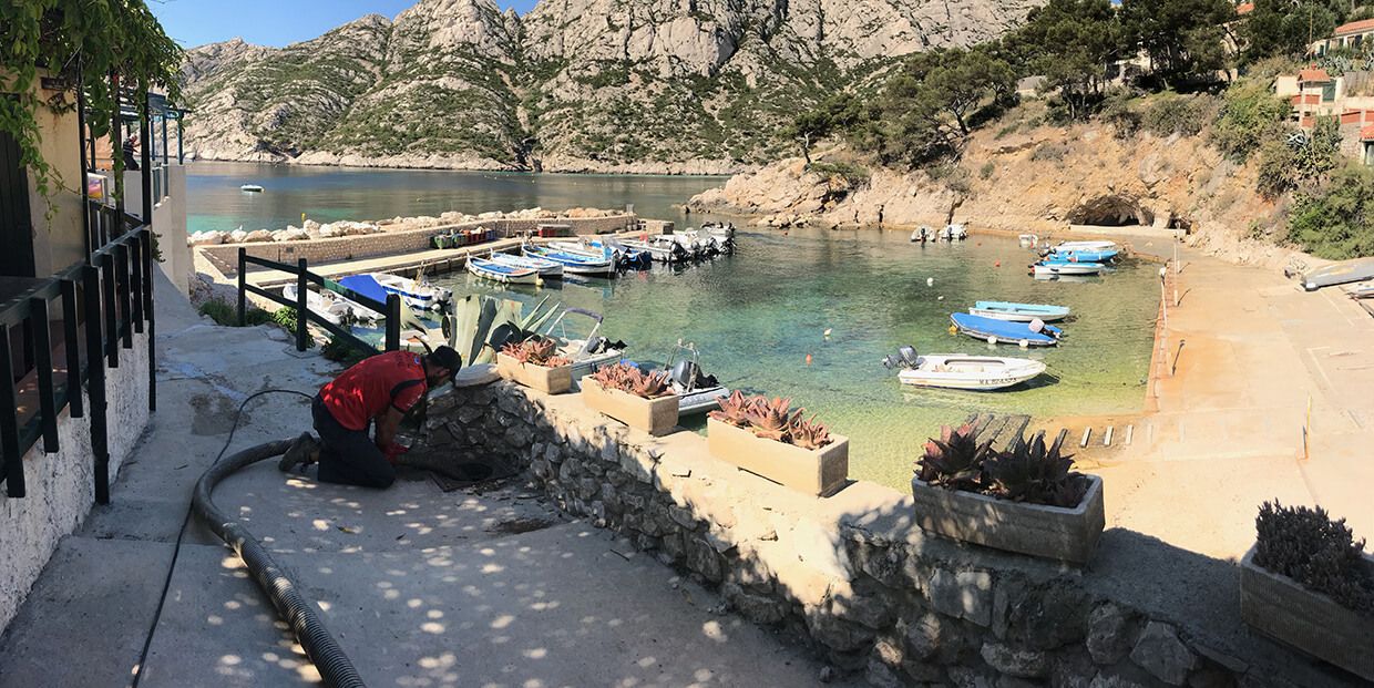 Petite crique aux eaux claires, bateaux amarrés et personnes sur une plage de sable fin bordant un chemin de pierres.