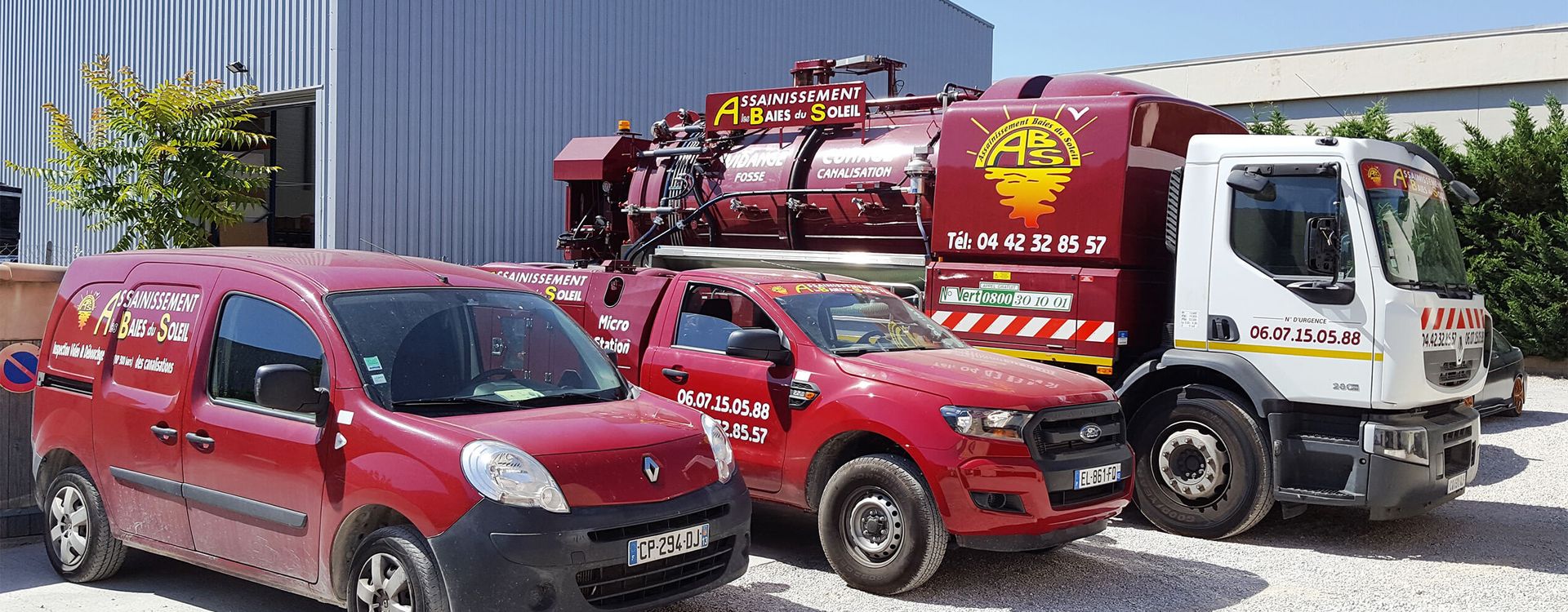 Un camion utilitaire rouge et deux fourgonnettes rouges sont garés à l'extérieur, sur du gravier, près d'un bâtiment en tôle ondulée.