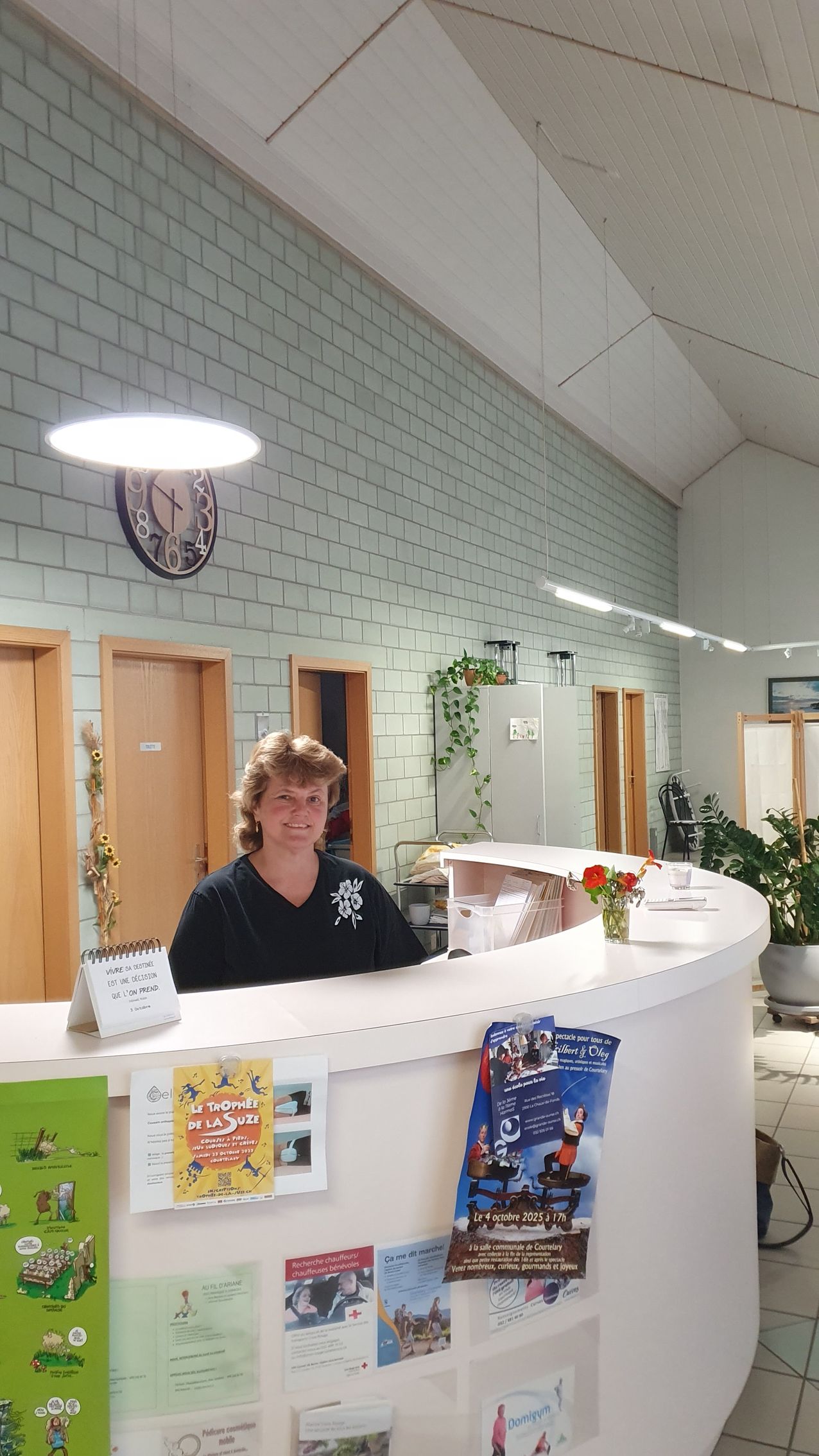 Un petit bâtiment gris avec une porte et des fenêtres roses, des fleurs en pot devant.