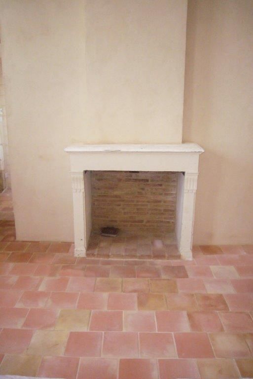 Cheminée vide avec manteau blanc dans une pièce beige et sol carrelé rose.