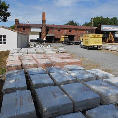Des palettes de marchandises emballées se trouvent dans une cour à l'extérieur d'un bâtiment industriel en briques, avec des camions et un chariot élévateur à proximité.