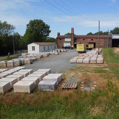 Des blocs de pierre empilés dans une cour industrielle, à côté d'un bâtiment en briques et d'un petit hangar blanc.