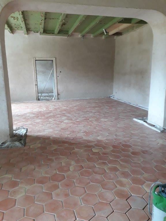 Pièce vide et inachevée avec sol en carrelage de terre cuite, murs beiges et poutres apparentes vertes au plafond.