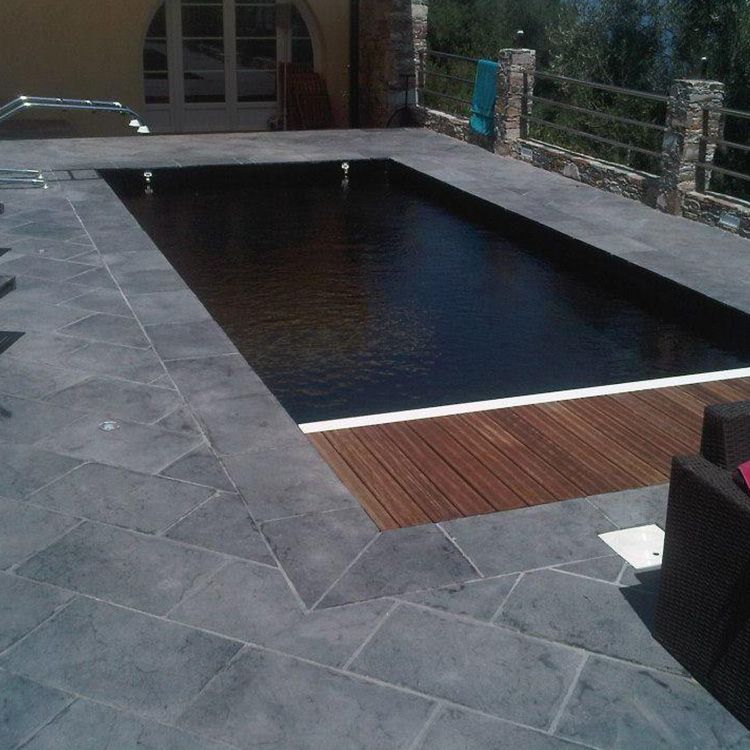 Piscine extérieure rectangulaire à l'eau sombre, terrasse en pierre grise et bordure en bois dans un cadre de jardin